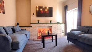 A living room with two sofas, a fireplace, a TV mounted above it, and a window with curtains. - 4 Bed • Parking • Near M4 • Ideal for Workers (Aberavon)