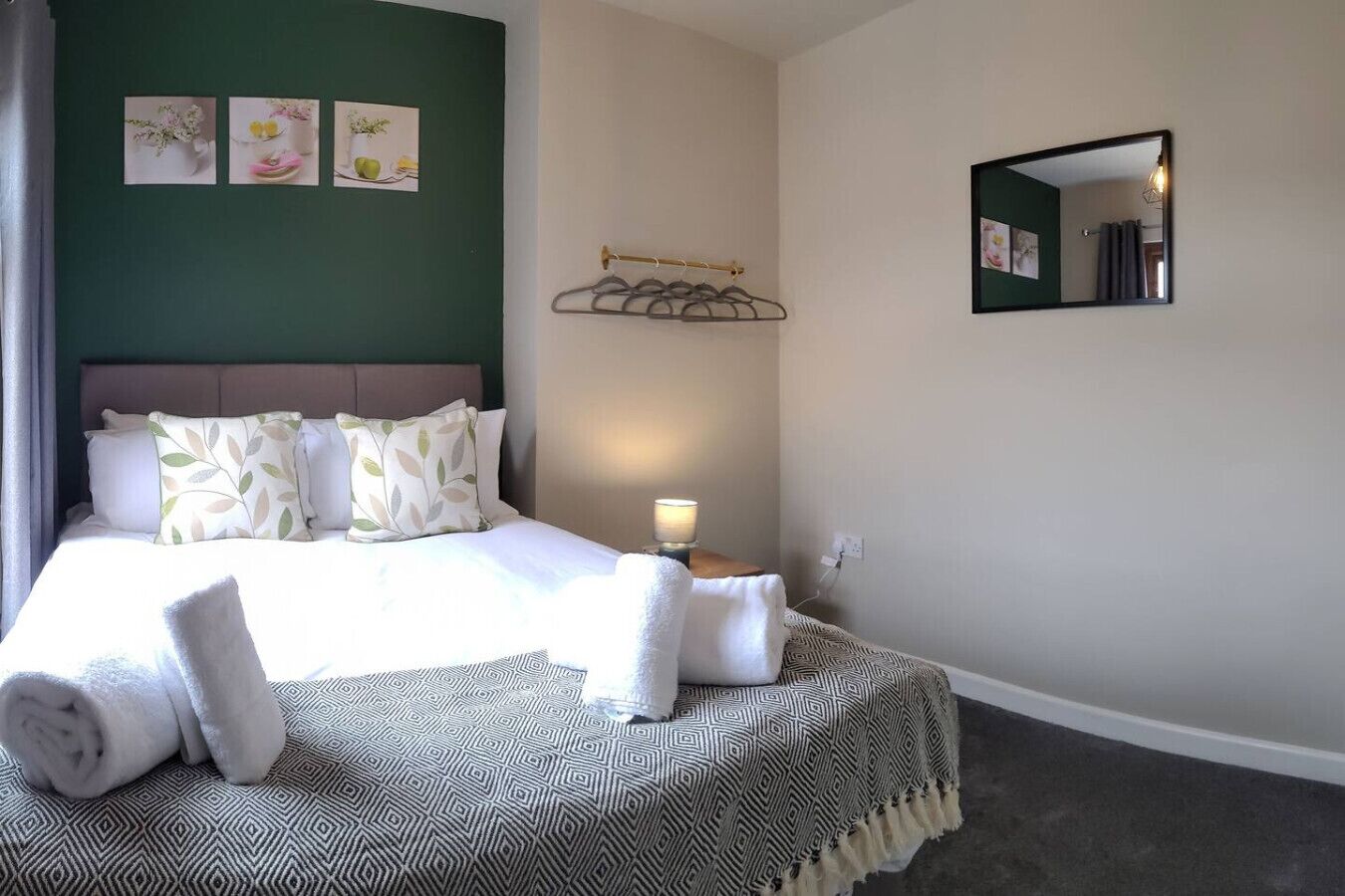A neatly made bed with pillows and a blanket, a bedside table with a lamp, a wall-mounted mirror, and a wall with three framed pictures.