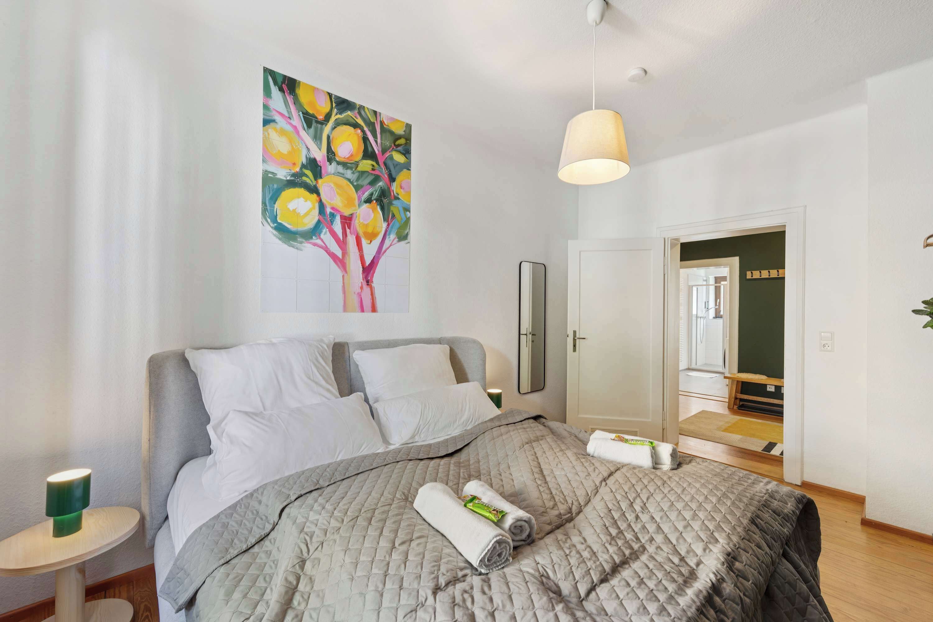Ein ordentlich bezogenes Bett mit gesteppter Decke, ein Baumgemälde mit Früchten, eine Nachttischlampe und ein Spiegel an der Wand.