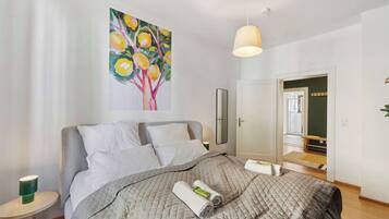 Ein ordentlich bezogenes Bett mit gesteppter Decke, ein Baumgemälde mit Früchten, eine Nachttischlampe und ein Spiegel an der Wand.