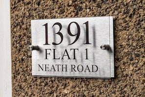 Metallinen talonumerokyltti, jossa lukee ”1391, ASUNTO 1, NEATH ROAD”, ja se on kiinnitetty kuvioituun seinään.