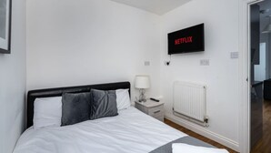 A modern bedroom with a bed, a bedside table with a lamp, a wall-mounted TV displaying 'NETFLIX,' and a white radiator.