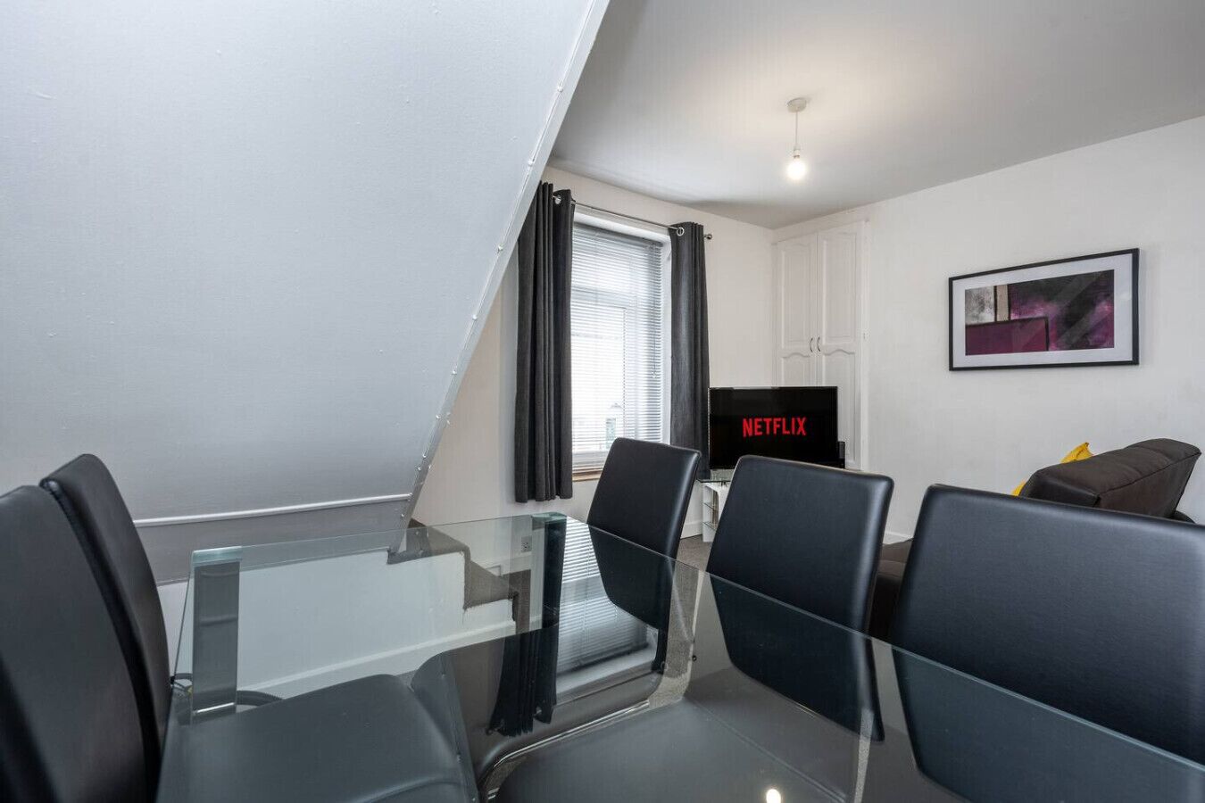 A modern living room with a glass table, black chairs, a flat-screen TV, and a framed picture on the wall.