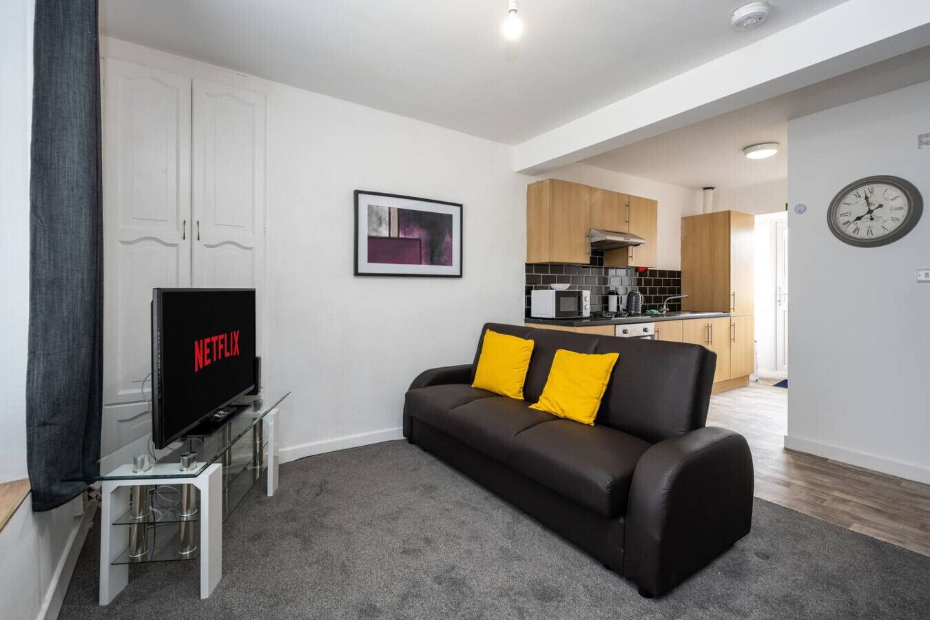 Una sala de estar moderna con un sofá de cuero negro, una mesa de centro de vidrio, un televisor de pantalla plana mostrando Netflix y una cocina al fondo.