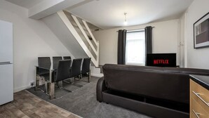 A modern living room with a sofa, a dining table with chairs, a staircase, a TV with Netflix logo, and a window with curtains. - King•En-suite Flat•Kitchen•Quiet Near Shops & Bus (Swansea)