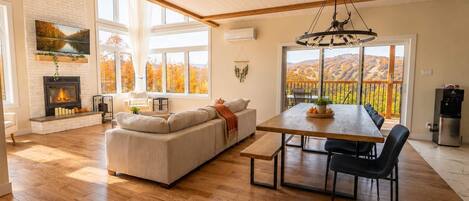 Ein geräumiges Wohnzimmer mit Kamin, einer großen Couch, einem Esstisch mit Stühlen und Blick auf herbstliche Laubfärbung durch große Fenster.