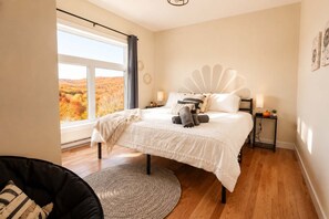 Ein Schlafzimmer mit großem Fenster, einem Bett mit weißer Bettwäsche, einem runden Teppich und einem Nachttisch mit Lampe.