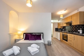 A hotel room with a bed, a bedside table with a lamp, a small trash bin, and a view into the kitchen area. - En-suite Flat • Kitchen • Quiet Near Shops & Bus (Swansea)