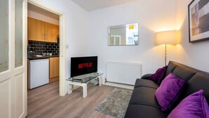Una sala de estar con televisión, sofá, lámpara, una cocina al fondo y un espejo en la pared.
