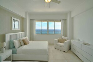 Une chambre d’hôtel avec un lit, une chaise, une commode, une fenêtre donnant sur la mer et un ventilateur de plafond.