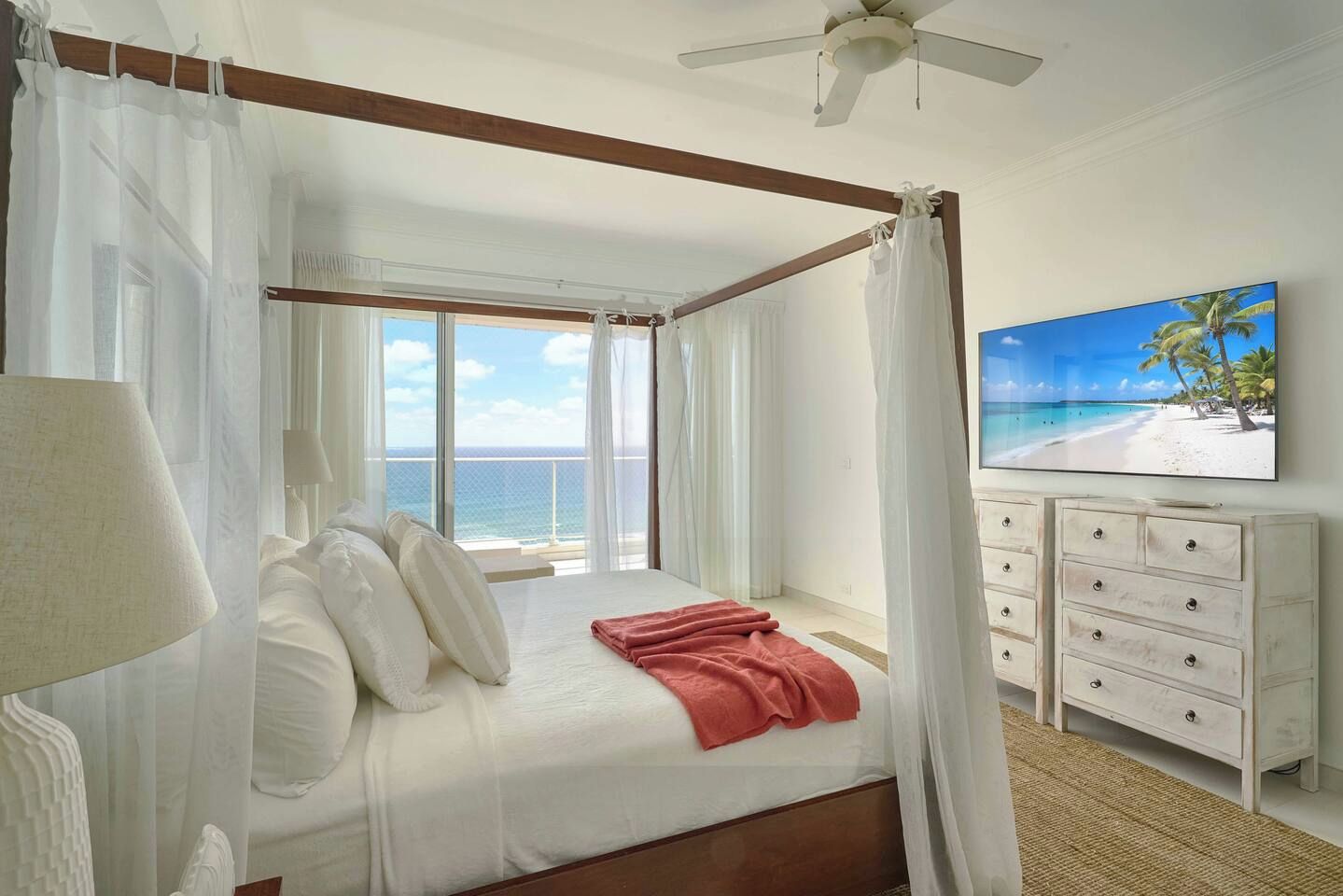 A four-poster bed with a view of the ocean, a ceiling fan, and a painting of a beach scene.