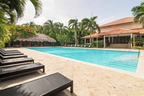 A swimming pool with lounge chairs, a thatched-roof structure, and a building with a tiled roof. - ®{3BR~Hemingway~Retreat}@JuanDolio+WalktoBeach+GYM (Juan Dolio)