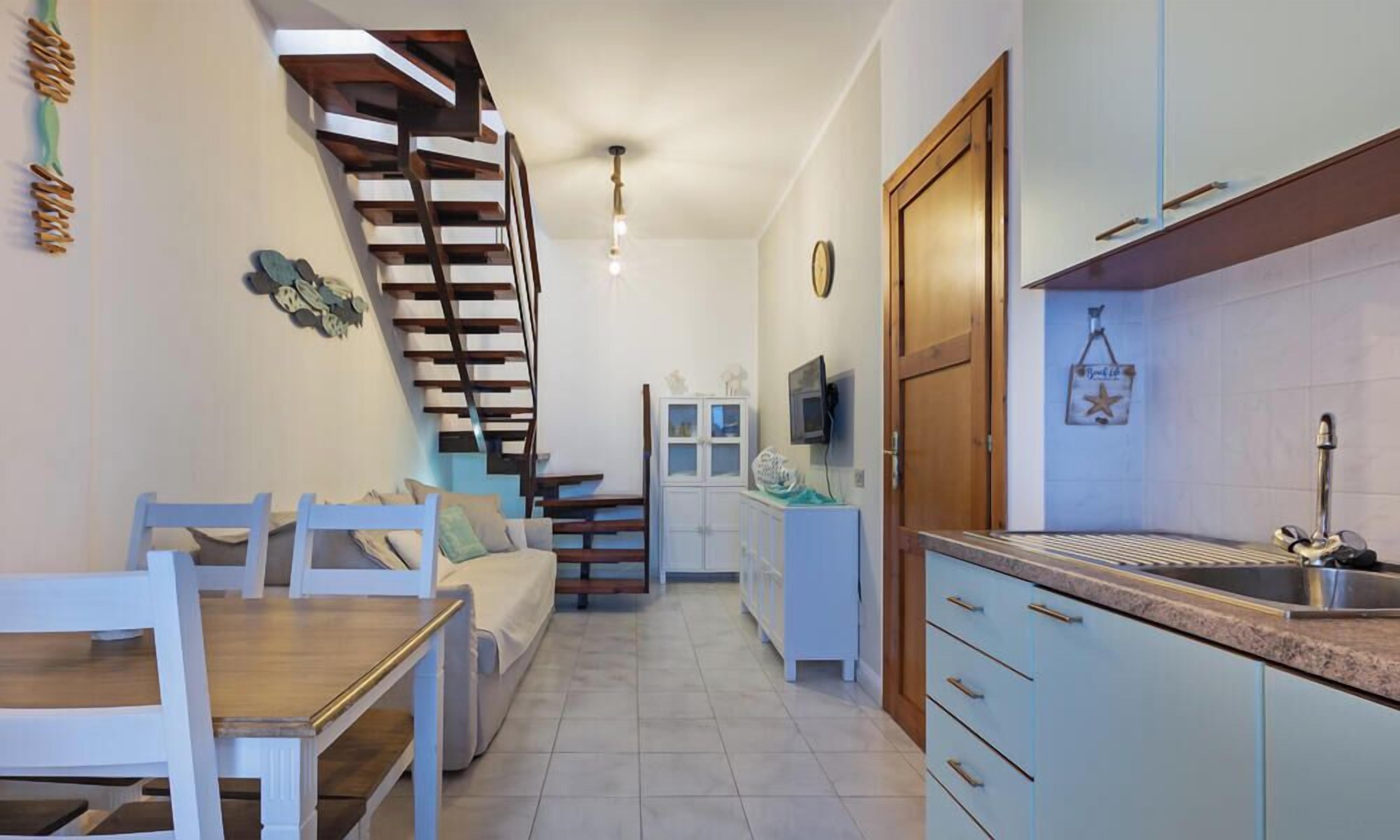 Una cocina moderna con estufa integrada, un comedor con mesa y sillas de madera, y una escalera que lleva a otro nivel.