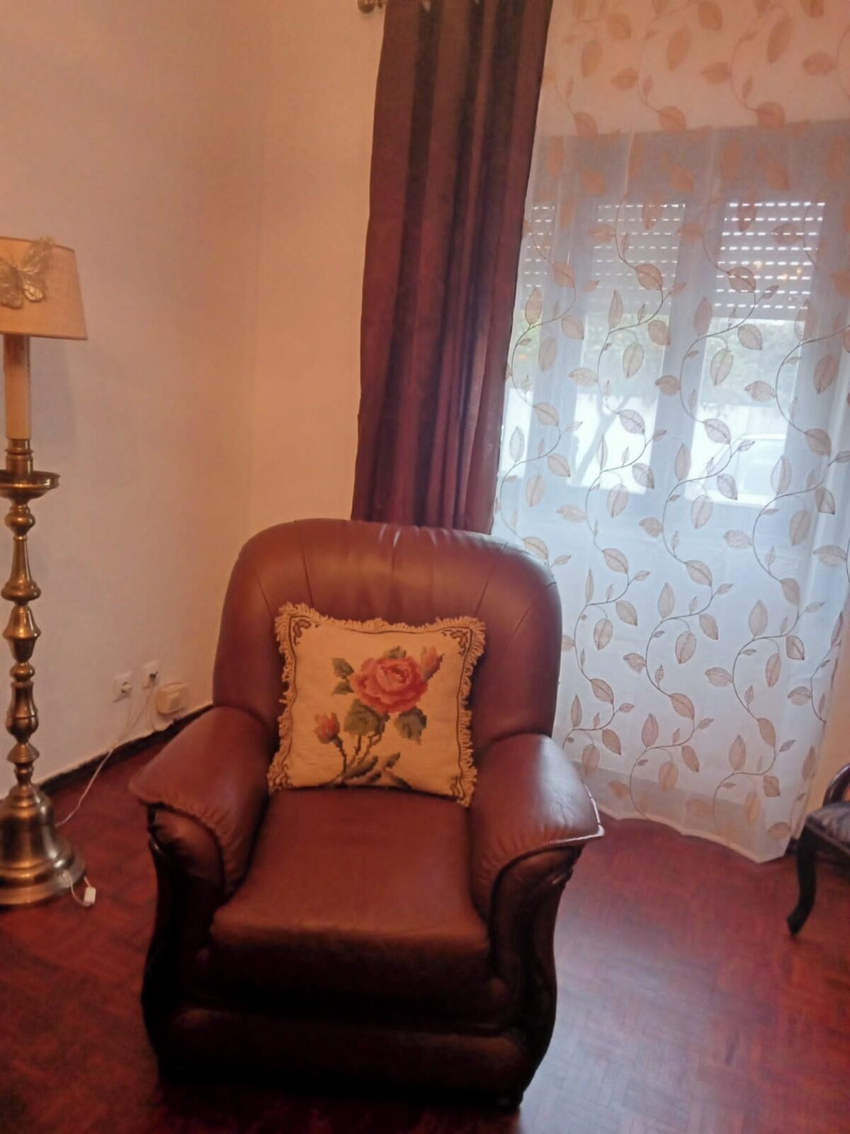 A brown leather armchair with a floral pillow, a lamp, and a window with sheer curtains.