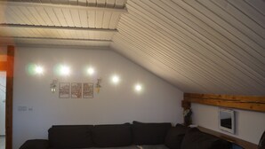 Ein Zimmer mit schrägem Dach, einem dunkelgrauen Sofa, Wandleuchten und einem kleinen gerahmten Bild.