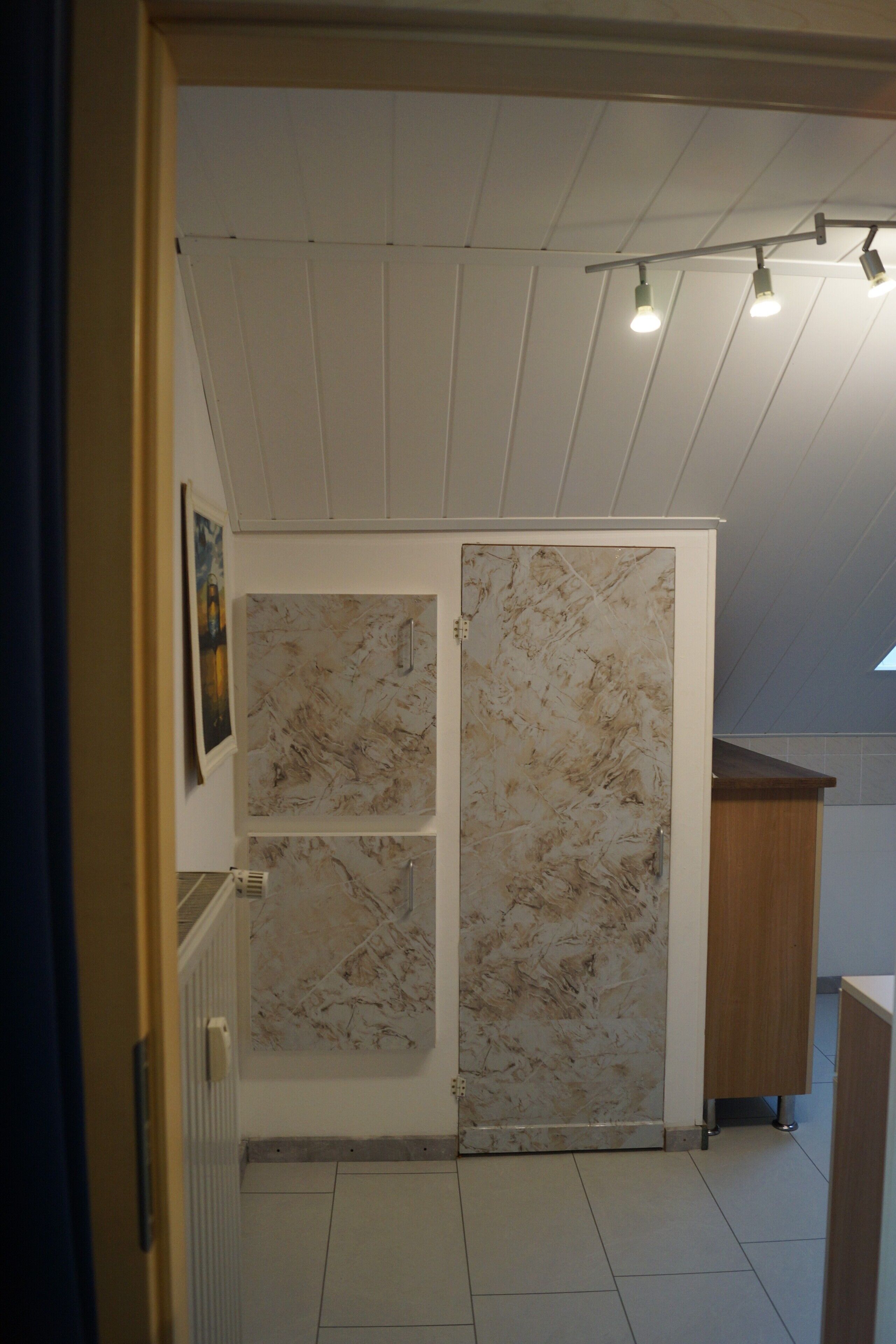Ein Innenraum mit Fliesenboden, einer Tür mit marmorartigem Verkleidungselement und einer Decke mit Einbaustrahlern.