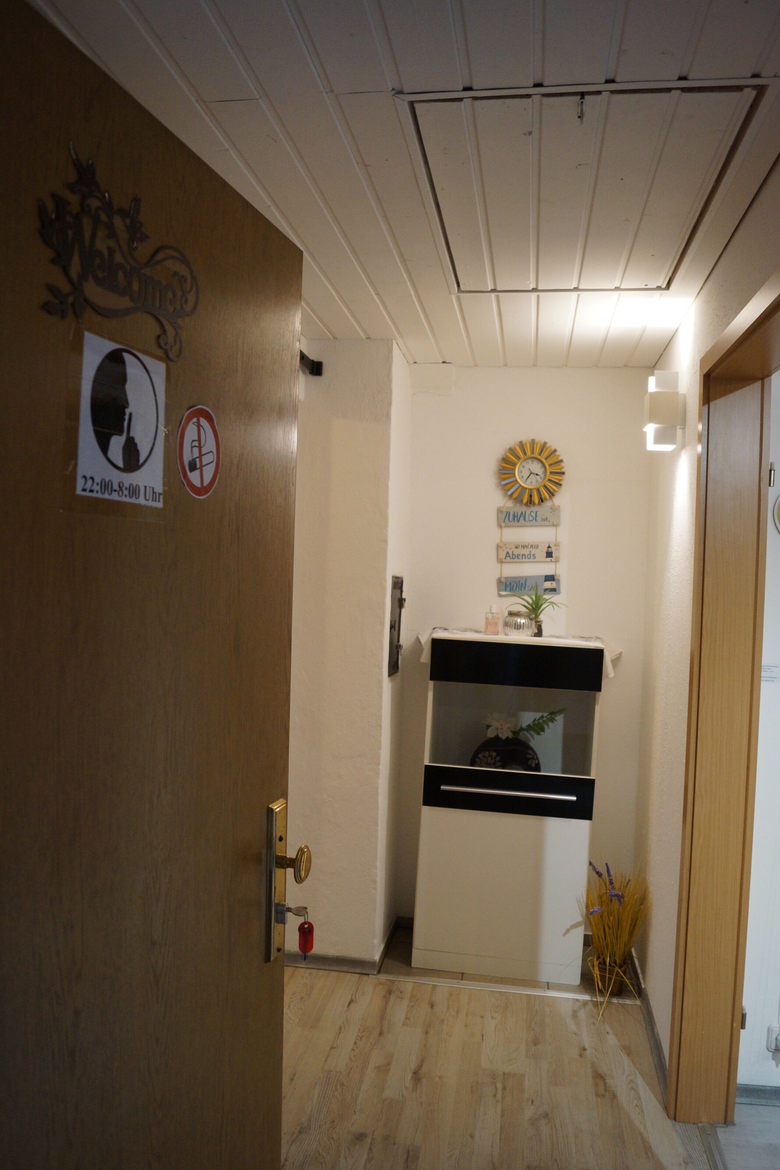 Een gang met een houten deur, een wandklok, een bord met een silhouet en een rookvrij-symbool, en een klein plankje met een vaas en een klok.