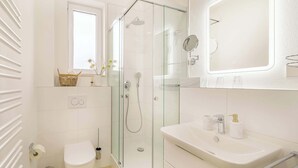Moderni kylpyhuone, jossa on lasinen suihkuseinä, wc-istuin, peilillä varustettu pesuallas ja ikkuna pesualtaan yläpuolella.