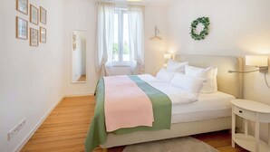 Ein Schlafzimmer mit Bett, Nachttischen, einer Pendelleuchte, einem Fenster mit Vorhängen, einem Spiegel und Wandbildern.