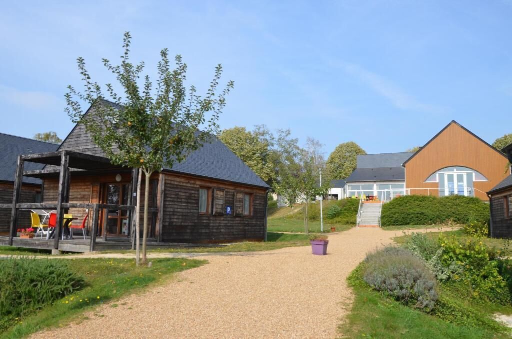 A wooden building with a covered outdoor seating area and a gravel path leading to a larger building with a sloped roof.