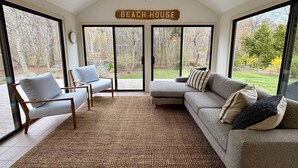 En rummelig opholdsstue med en stor skydedør til en have, en ventilator i loftet, og et skilt med teksten 'BEACH HOUSE'.