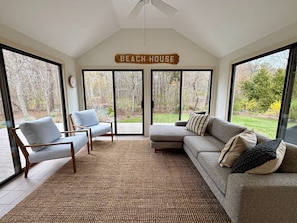 A spacious living area with a large sliding glass door leading to a garden, a ceiling fan, and a sign reading 'BEACH HOUSE'.