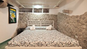 Une chambre avec un mur en pierre, un lit recouvert d’une literie à motifs, un tableau accroché au mur et une étagère ornée d’objets décoratifs.