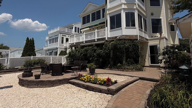 A multi-story house with a balcony, a patio area with outdoor furniture, and a garden with flowers. - Lagoon Luxe at Osborn Island - Private Dock - Minutes to LBI and Graveling Beach (Little Egg Harbor Township)
