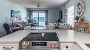 Cozinha moderna com ilha central, área de jantar com mesa e cadeiras, sala de estar com sofá e cadeiras, e varanda com vista.