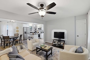 A modern living room with a ceiling fan, a dining area with a table and chairs, a kitchen in the background, and a television on a stand.