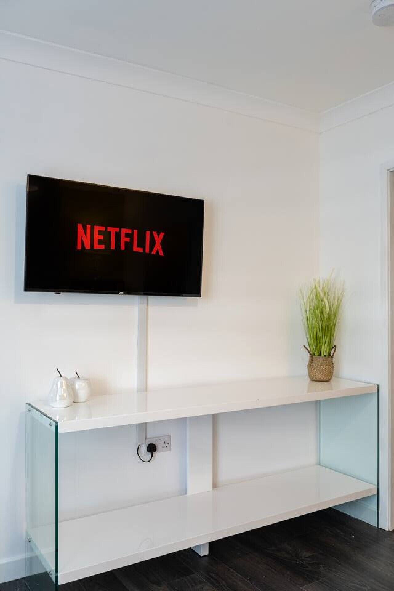 A modern living room with a mounted TV displaying 'NETFLIX,' a glass console table, a potted plant, and a power outlet.