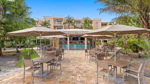 Un patio al aire libre con mesas y sombrillas, piscina y un edificio con balcones.