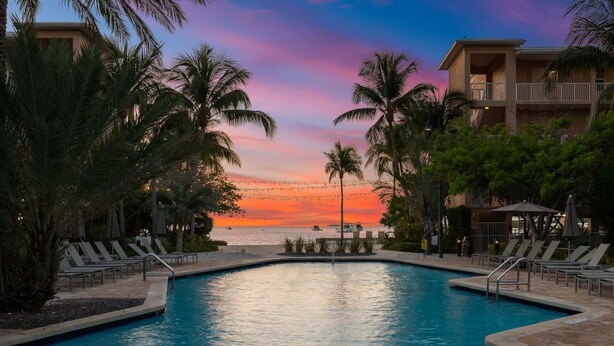 Una piscina con sillas de descanso, palmeras y vistas al atardecer sobre el agua.