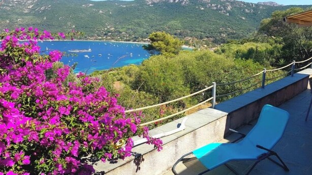 Un balcón con vistas a la costa, una silla y flores moradas.