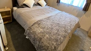 A single bed with a dark headboard, a bedside table, a window with curtains, and a carpeted floor. - Elegant 2 Bed One and Half Bath Caravan (Northampton)