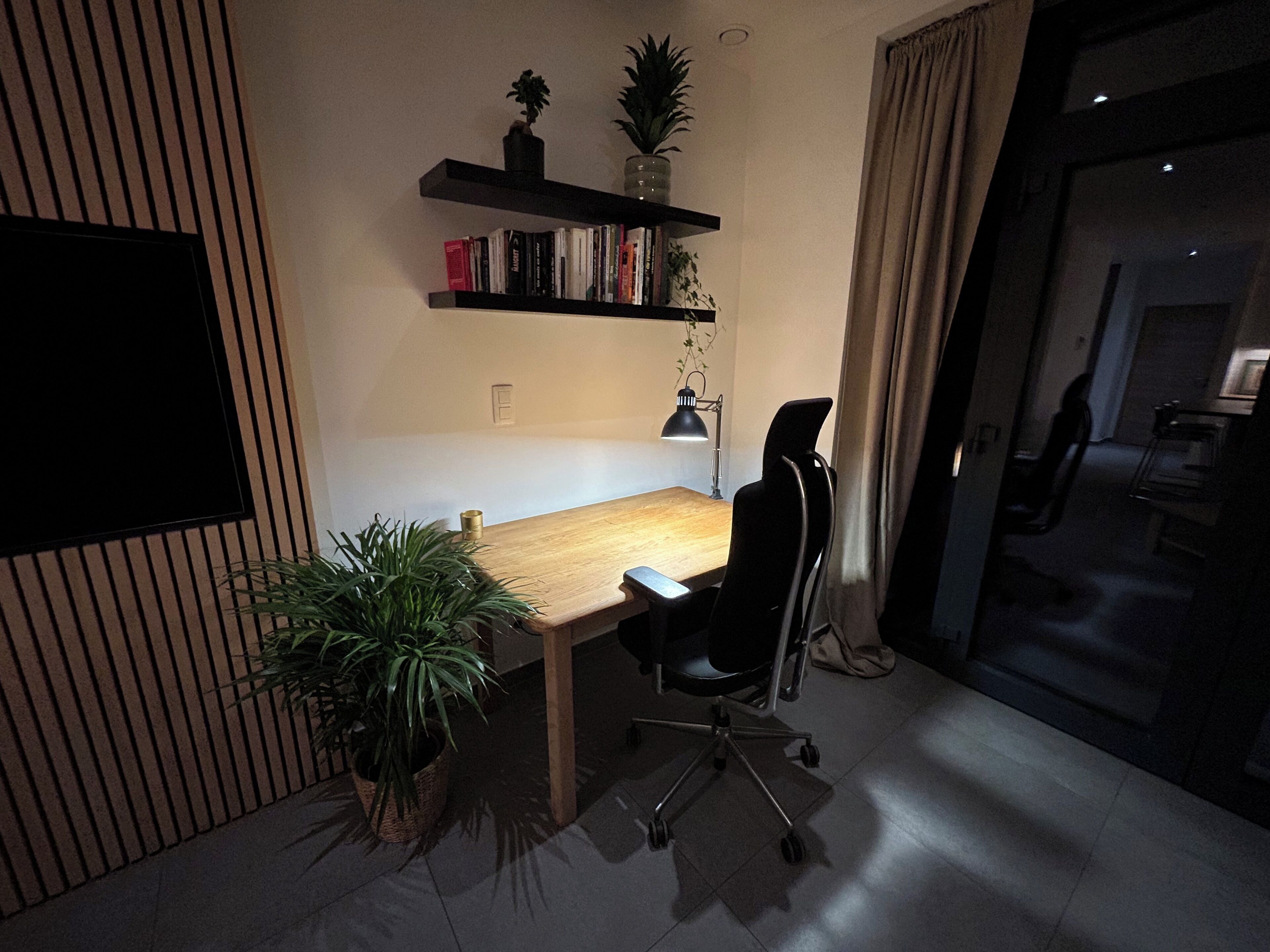 A room with a wooden desk, a chair, a TV mounted on the wall, a bookshelf with books and plants, and a window with curtains.