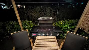 Un área de parrilla al aire libre con una mesa y dos sillas, rodeada de follaje.