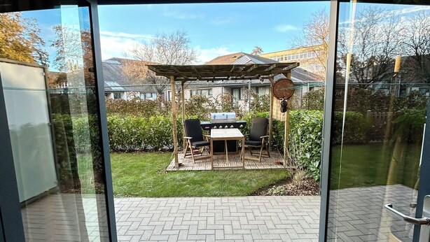 Una puerta de cristal moderna que da a un patio con una parrilla, mobiliario exterior y vistas del entorno.