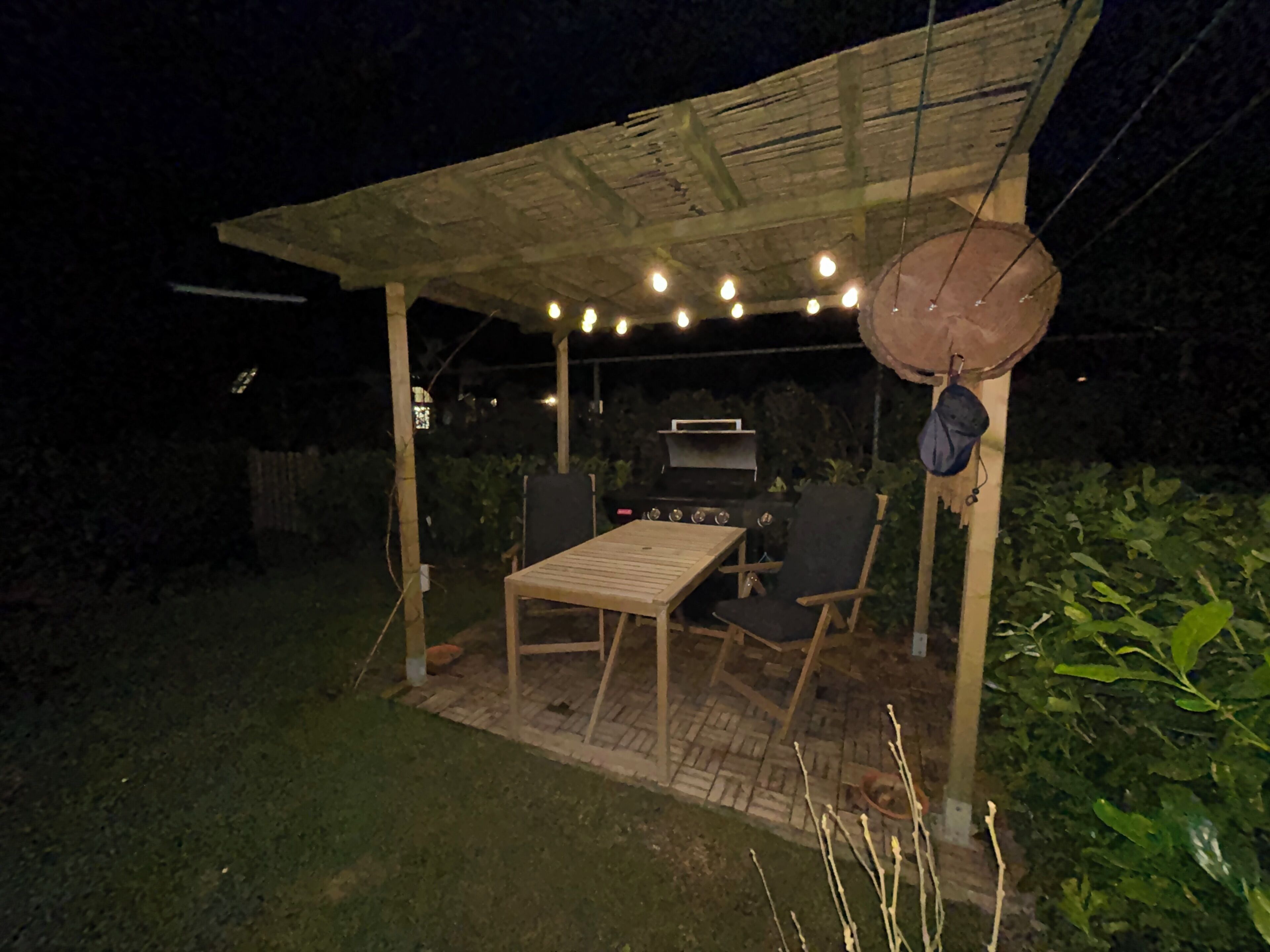 Une terrasse extérieure avec une table et des chaises en bois, un barbecue, des guirlandes lumineuses et un toit de chaume.