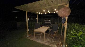 Une terrasse extérieure avec une table et des chaises en bois, un barbecue, des guirlandes lumineuses et un toit de chaume.