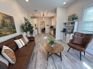 A modern living room with a brown sofa, a wooden coffee table, and a leather armchair. There is a kitchen in the background with white cabinets and a dining area.
