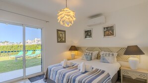 Ett sovrum med en säng, två sängbord, en hängande lampa och utsikt över en grön gräsmatta och byggnader.