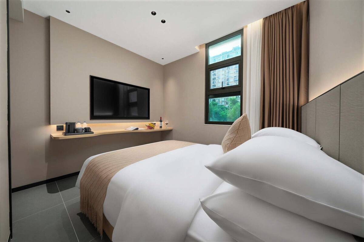 Una habitación de hotel moderna con una cama, un televisor montado en la pared, una ventana con cortinas y una mesita de noche con una lámpara y una taza.