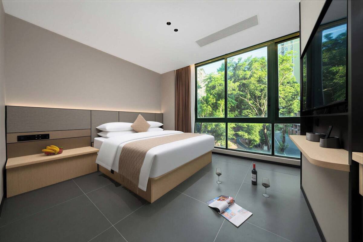 Una habitación de hotel moderna con una cama grande, una mesita de noche con frutero, una revista y una botella de vino.