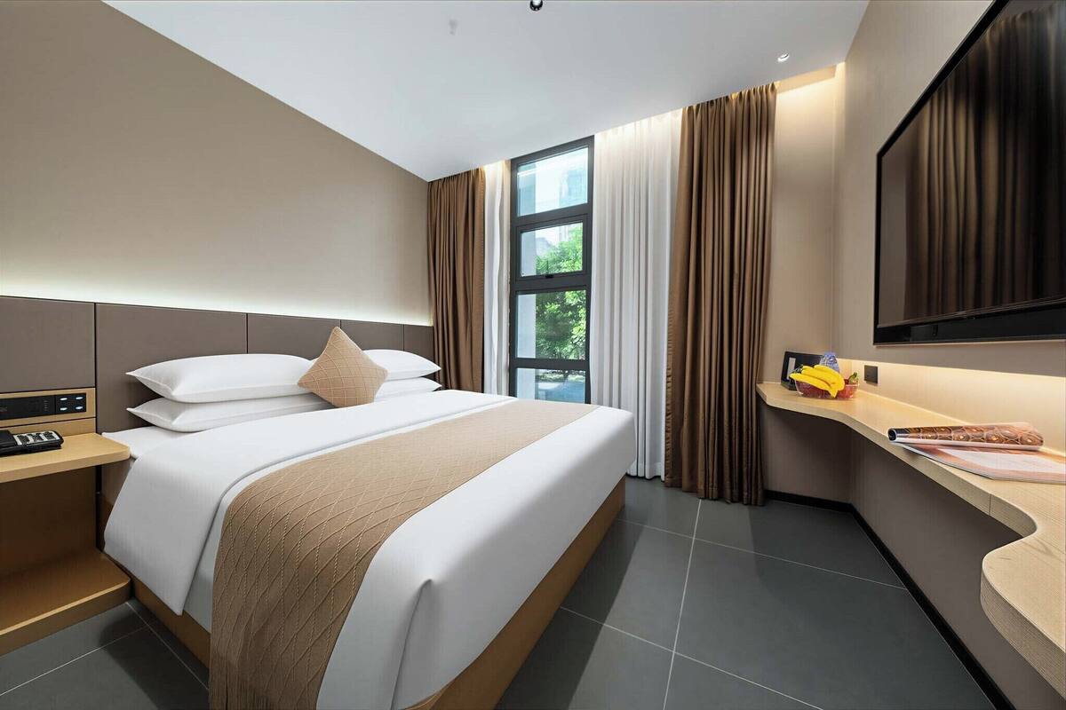 Una habitación de hotel moderna con cama, mesita de noche, televisor y ventana con cortinas.