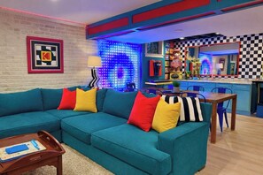 A teal sofa with colorful cushions, a wooden coffee table, a checkered wall, and a neon-lit wall.