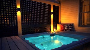 A hot tub with a wooden deck, a lattice wall, and a window with a cushion.