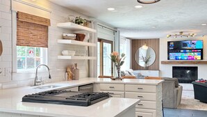A modern kitchen with white cabinetry, a built-in gas stove, and a large island. There is a dining area with a sofa and a TV mounted above a fireplace.