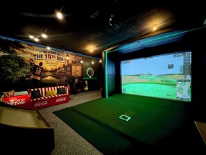 Une pièce dotée d’un grand écran diffusant un match de golf, d’un mur orné d’une pancarte indiquant « BIENVENUE AU 19E TROU », et d’un canapé à l’effigie de Coca-Cola.