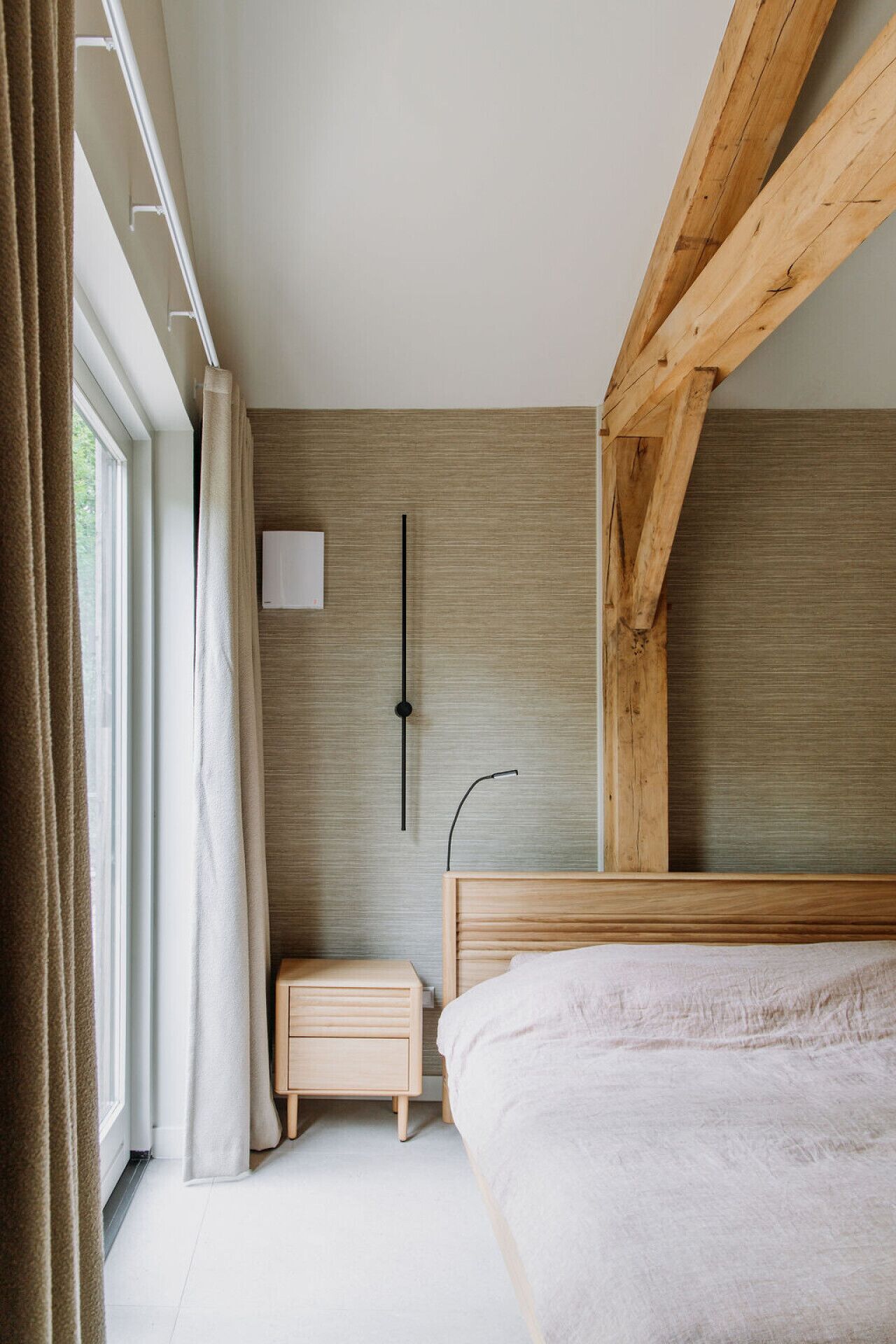 A bedroom with a wooden headboard, a bedside table, a nightstand, a wall-mounted light, and a window with curtains.